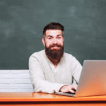 man sitting at a table typing on a laptop in front of a chalkboard