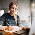 woman writing in a notebook while using a laptop