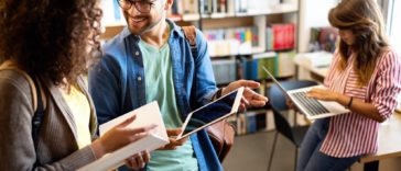 group of students in a library looking at a book, a laptop and a tablet