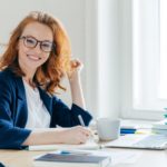 smiling woman sitting at a table in front of a laptop and notebook holding a pen