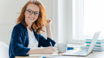 smiling woman sitting at a table in front of a laptop and notebook holding a pen