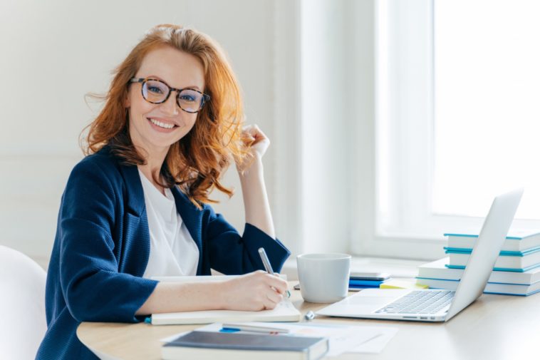 smiling woman sitting at a table in front of a laptop and notebook holding a pen