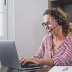 woman sitting at a work table typing on a laptop and talking on headphones