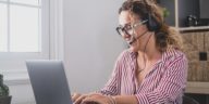 woman sitting at a work table typing on a laptop and talking on headphones