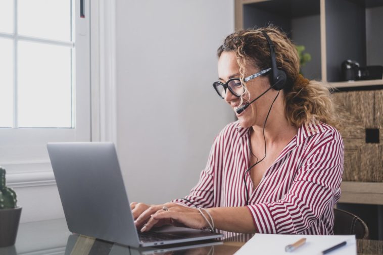 woman sitting at a work table typing on a laptop and talking on headphones