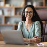 smiling woman with headphones sitting behind a desk with a laptop