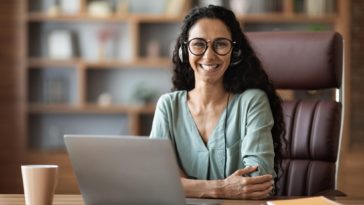 smiling woman with headphones sitting behind a desk with a laptop