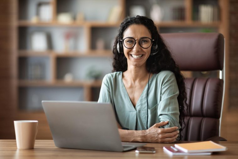 smiling woman with headphones sitting behind a desk with a laptop