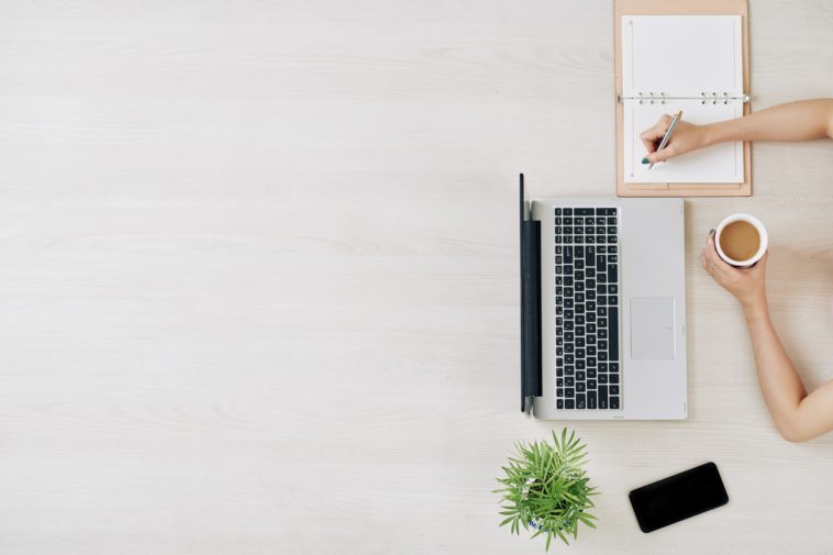 bird view of laptop, phone, plant, coffee and someone writing in a notebook