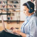 woman with headphones sitting on the floor of a library and typing on a laptop