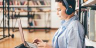 woman with headphones sitting on the floor of a library and typing on a laptop