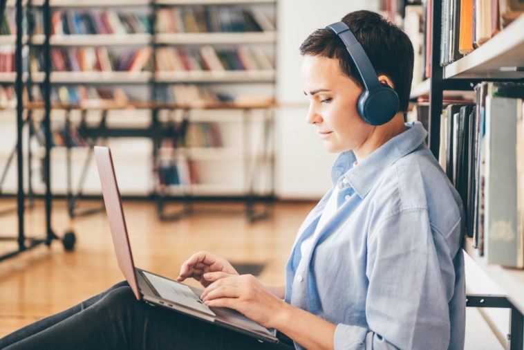 woman with headphones sitting on the floor of a library and typing on a laptop