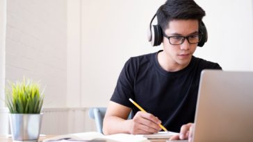 young man with headphones looking at a laptop and writing