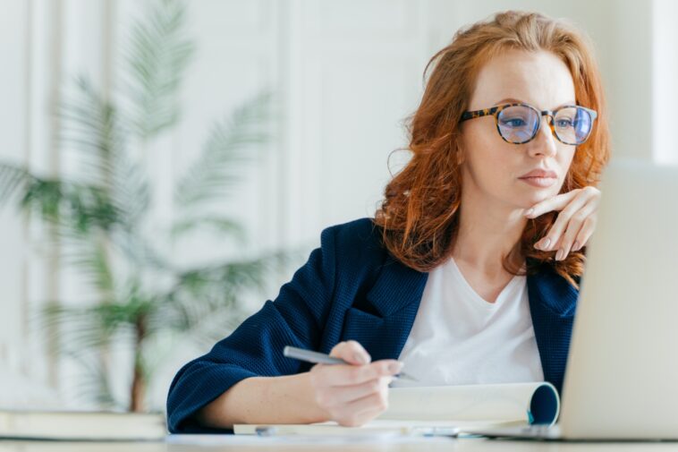 woman looking at a computer making notes in a notepad