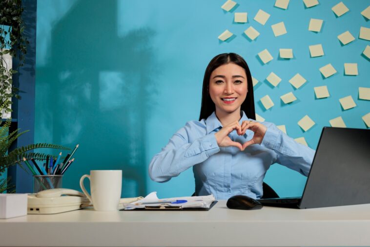 woman making a heart shaped sign sitting at a desk in a home office