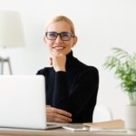 smiling woman with glasses sitting at a desk with a laptop