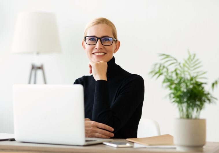 smiling woman with glasses sitting at a desk with a laptop