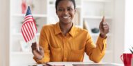 smiling woman sitting at a desk with a laptop holding a flag of the usa