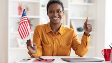 smiling woman sitting at a desk with a laptop holding a flag of the usa