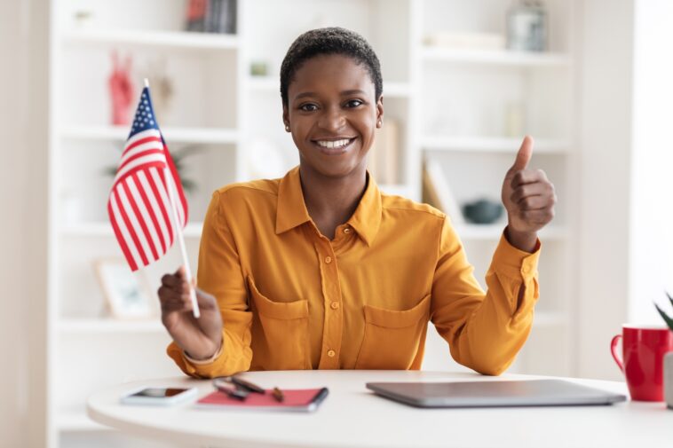smiling woman sitting at a desk with a laptop holding a flag of the usa