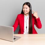 woman sitting behind a desk with a laptop pointing at her headset