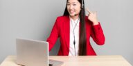 woman sitting behind a desk with a laptop pointing at her headset