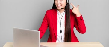 woman sitting behind a desk with a laptop pointing at her headset