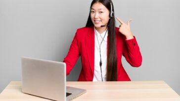 woman sitting behind a desk with a laptop pointing at her headset