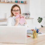 woman sitting at a desk, holding a notebook and looking at a laptop