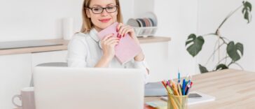 woman sitting at a desk, holding a notebook and looking at a laptop