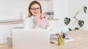 woman sitting at a desk, holding a notebook and looking at a laptop