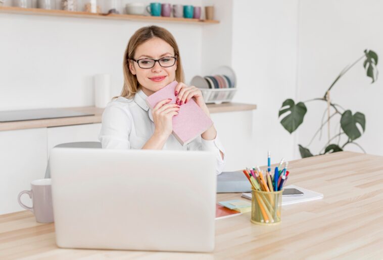 woman sitting at a desk, holding a notebook and looking at a laptop