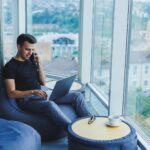 man sitting in a modern home office using a laptop and talking on a phone