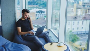 man sitting in a modern home office using a laptop and talking on a phone