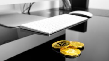 3 gold bitcoins on a black table next to a white computer