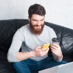 man sitting on a sofa using a smartphone and holding a coffee cup