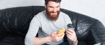 man sitting on a sofa using a smartphone and holding a coffee cup