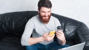 man sitting on a sofa using a smartphone and holding a coffee cup