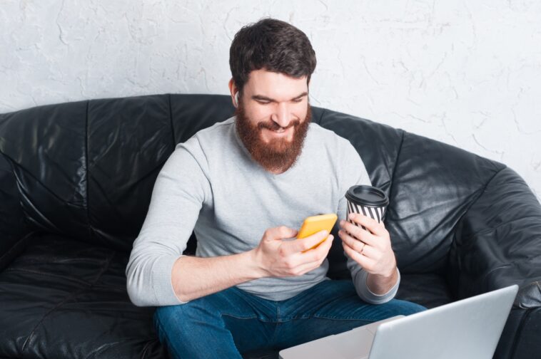 man sitting on a sofa using a smartphone and holding a coffee cup