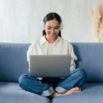 smiley woman with headphones sitting on a couch and working on a laptop