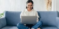 smiley woman with headphones sitting on a couch and working on a laptop