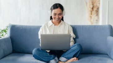 smiley woman with headphones sitting on a couch and working on a laptop