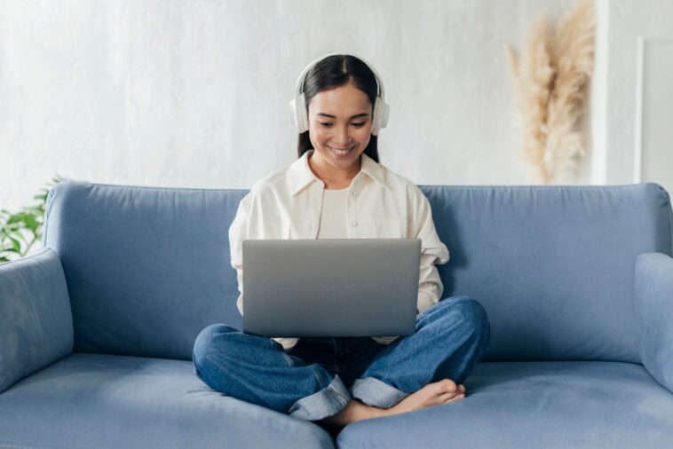 smiley woman with headphones sitting on a couch and working on a laptop